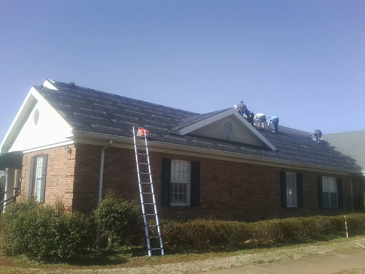 Crew tearing shingles off a brick ranch roof with a ladder set — Metal Roof Repair work by our crew in Cornville