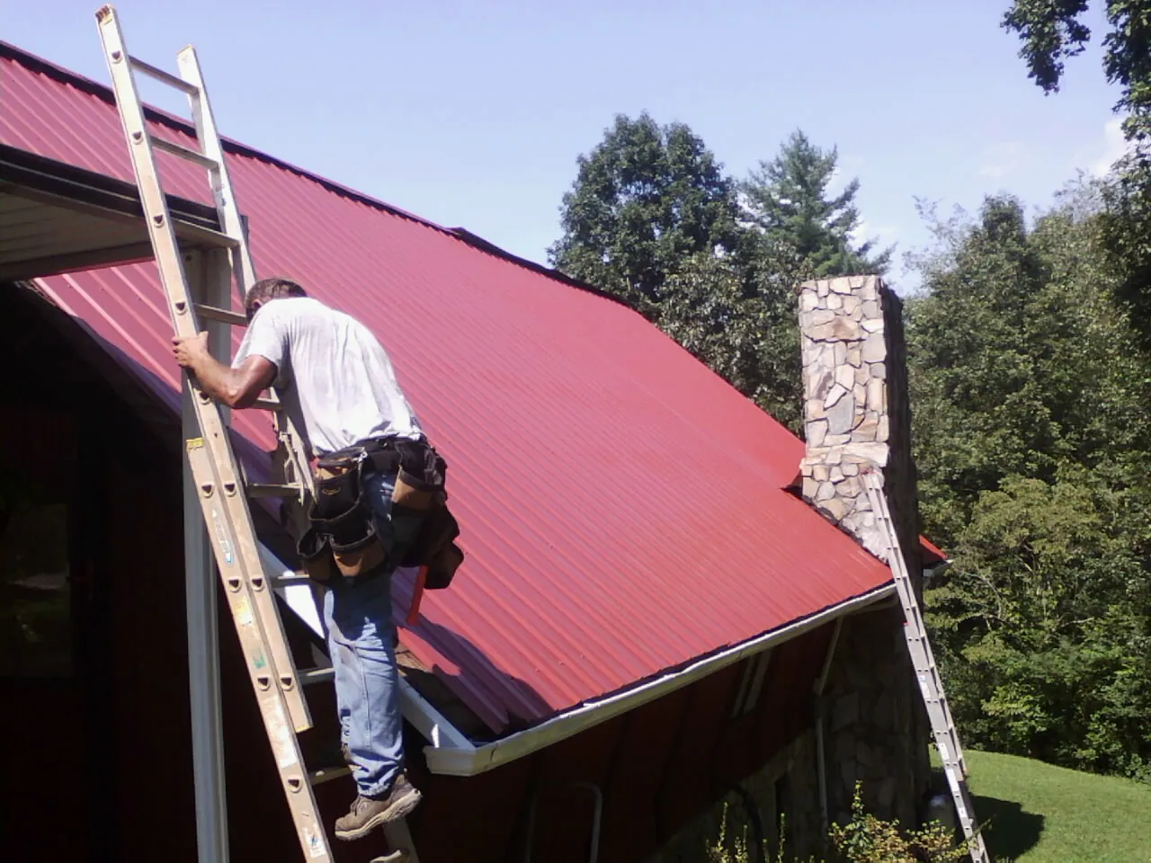 Roofer on a ladder beside a red metal roof and stone chimney in Cornville